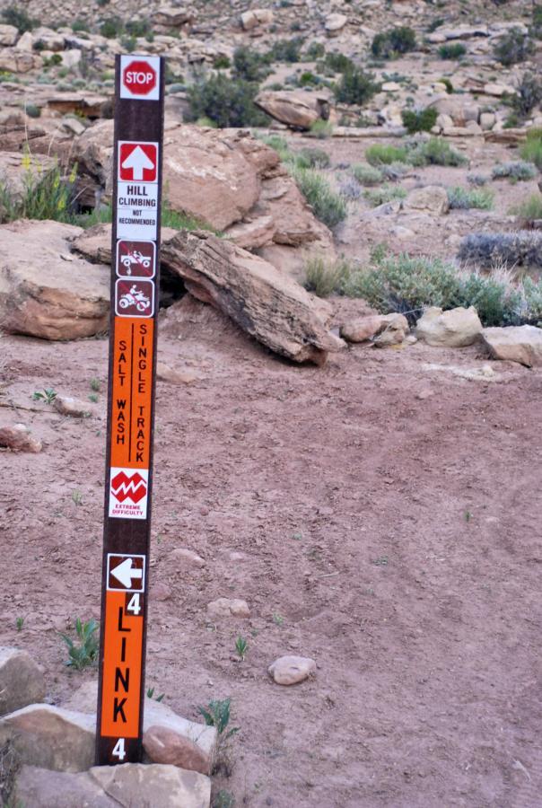 A trail marker sign in a rocky outdoor landscape, featuring multiple directional arrows and information. The sign includes a "STOP" symbol, a recommendation against hill climbing, and indicates trails such as "Salt Wash" and "Link 4" with difficulty levels. The background consists of rocky terrain and sparse vegetation. Sovereign Single Track mountain bike trail.