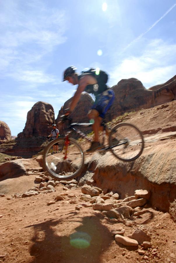 A mountain biker jumps over a rocky section of trail with red rock formations and a clear blue sky in the background. Another person is visible in the distance, observing the action. Amasa Back Trail mountain bike trail.
