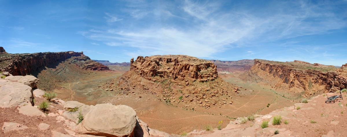 A panoramic view of a rugged desert landscape featuring dramatic rock formations and a clear blue sky. The scene showcases a wide valley surrounded by steep cliffs and mesas, highlighting the natural beauty and geology of the area. In the foreground, there are scattered rocks and patches of greenery, while the background reveals distant mountains and a vast open sky. Amasa Back Trail mountain bike trail.