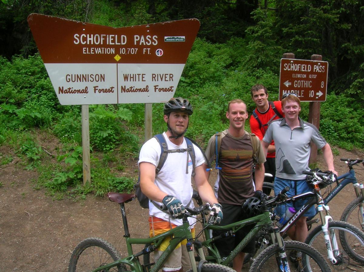 Four mountain bikers pose with their bikes in front of a sign marking Schofield Pass, which has an elevation of 10,707 feet. The background features lush green foliage typical of a national forest. Trail 401 mountain bike trail.