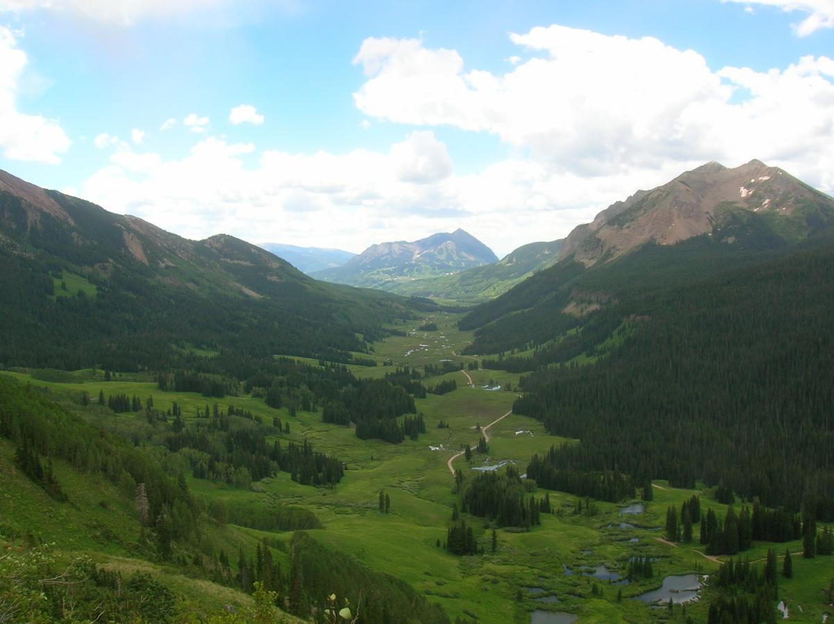 A panoramic view of a lush green valley surrounded by mountains under a partly cloudy sky. The landscape features dense forests, meadows, and winding roads, with small bodies of water scattered throughout the valley floor. Trail 401 mountain bike trail.