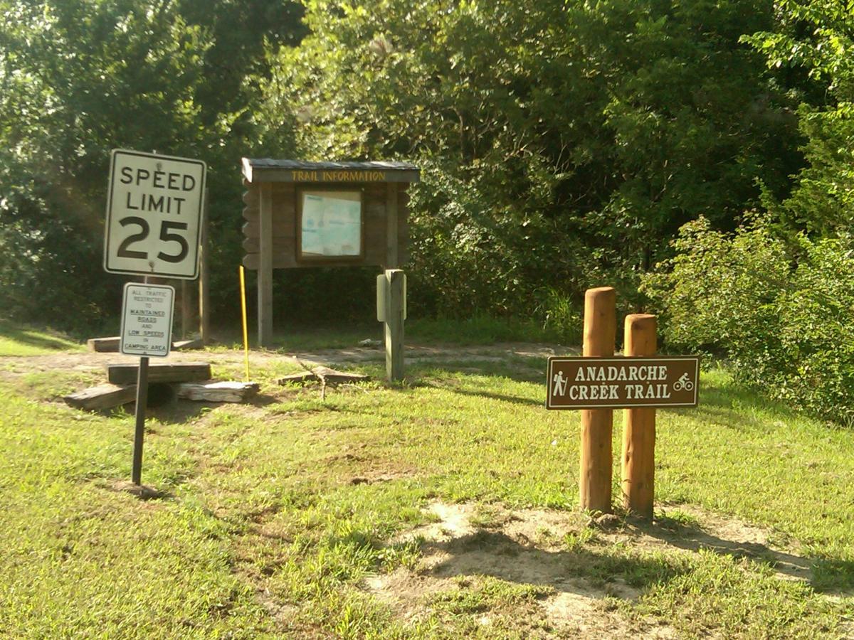 A grassy area featuring a speed limit sign indicating 25 mph, a wooden trail information kiosk, and a sign marking the beginning of the Anadarche Creek Trail, surrounded by trees and shrubbery. Lake Murray State Park mountain bike trail.