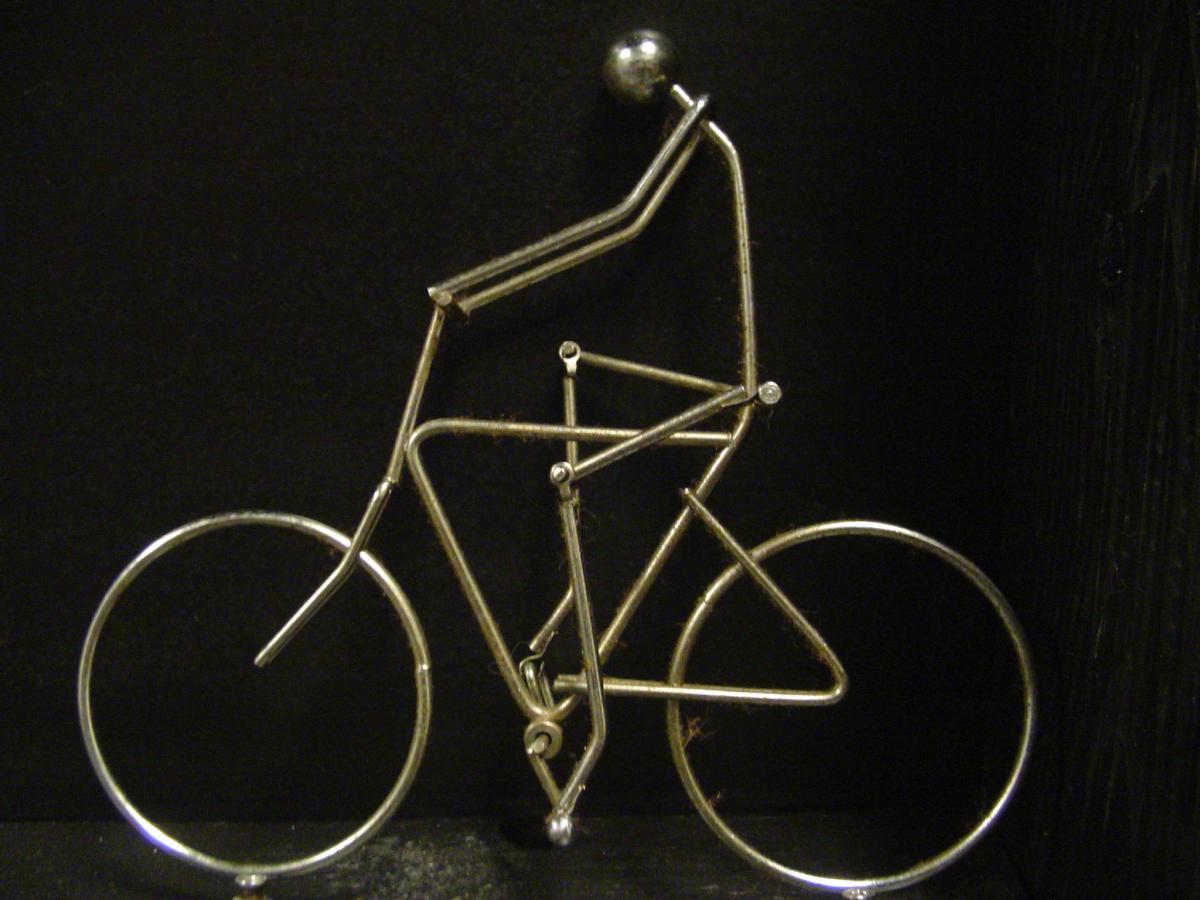A metallic sculpture of a bicycle with a stick figure rider, crafted from thin metal wire. The figure is in a cycling position, and both wheels are circular, creating a minimalist design against a black background.