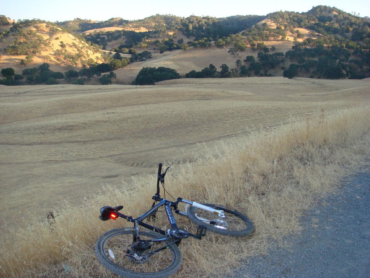 A black mountain bike rests on the ground beside a gravel path, surrounded by dry grass and rolling hills in the background. The landscape is sunlit, showcasing a mix of light and shadow across the golden terrain and a few scattered trees. Black Diamond Mines mountain bike trail.