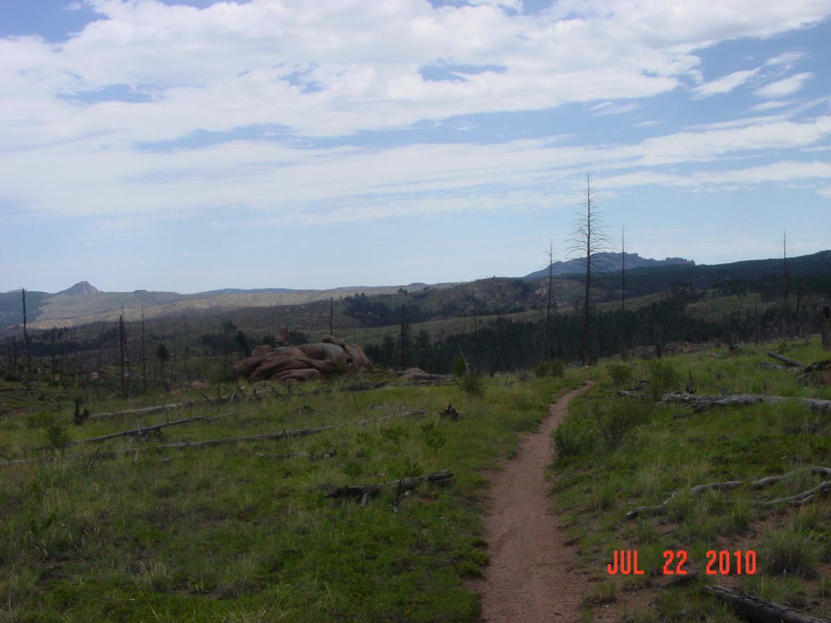 A winding dirt path traverses a grassy landscape, flanked by rocky outcrops and sparse trees. In the background, rolling hills rise under a partly cloudy sky, suggesting a serene natural setting. The scene captures the beauty of an expansive outdoor area with a mix of greenery and rocky terrain. Buffalo Creek mountain bike trail.