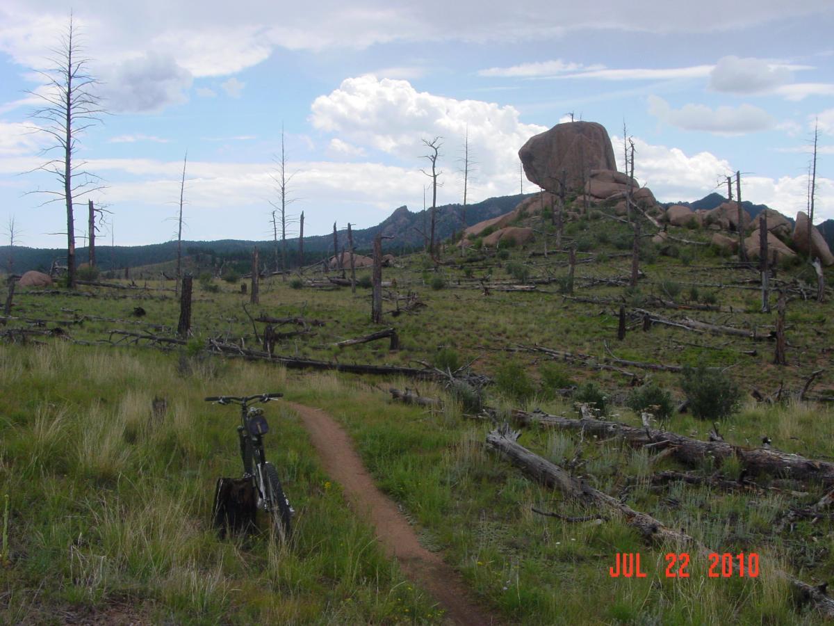 A mountain biking trail in a forested area with sparse vegetation, showing a bicycle leaning against a tree stump. In the background, large boulders and rocky outcrops rise above the landscape, with dead trees scattered throughout the scene under a cloudy sky. The image captures a moment in nature on July 22, 2010. Buffalo Creek mountain bike trail.