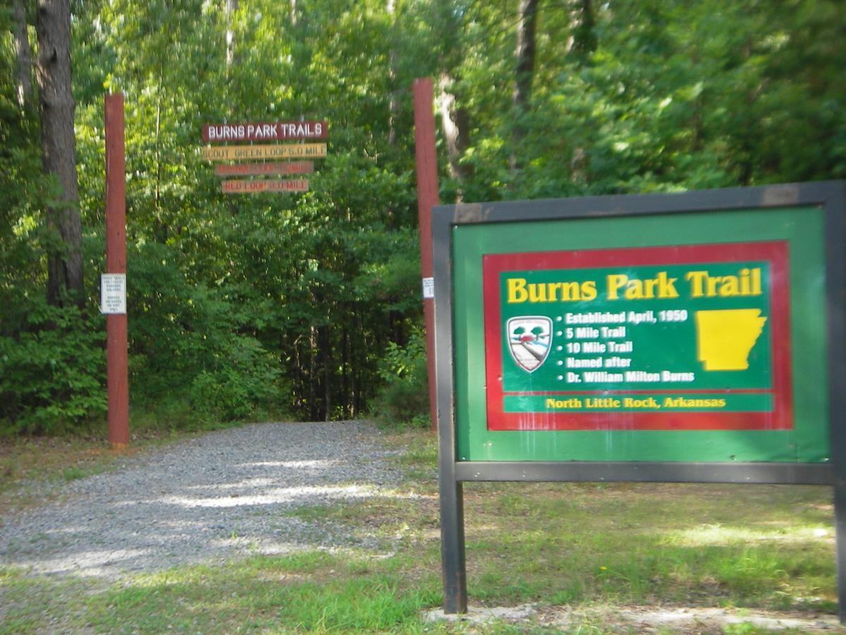 A wooden sign for Burns Park Trails in North Little Rock, Arkansas, indicating the Scout Green Loop (0.5 mile), a 5-mile trail, and a 10-mile trail. The sign also mentions that the trails were established in April 1950 and are named after Dr. William Milton Burns. Surrounding the sign is a forested area with green trees and a gravel path leading into the trails. Burns Park mountain bike trail.