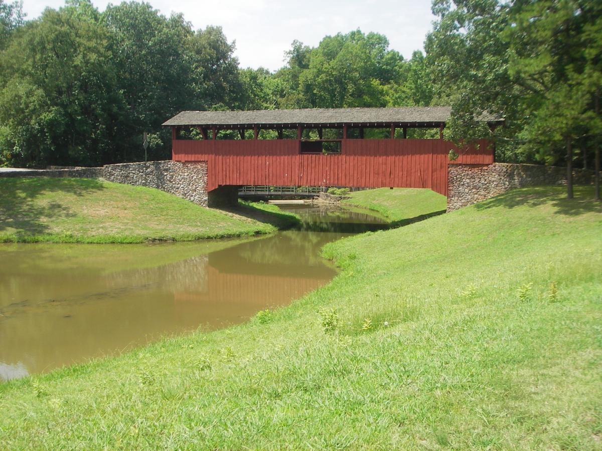 A picturesque scene of a red covered bridge spanning over a calm pond, surrounded by lush greenery and trees under a bright sky. The bridge features a stone foundation and a rustic wooden structure, reflecting in the water below. Gradually sloping grassy banks lead up to the bridge, enhancing the tranquil setting. Burns Park mountain bike trail.