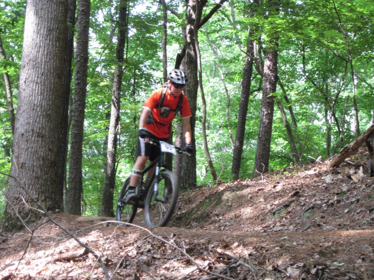 A young man wearing an orange shirt and a helmet rides a mountain bike on a dirt trail surrounded by green trees and foliage. The terrain is slightly uneven, with leaves and branches scattered along the path. Oak Mountain State Park Bump Trail mountain bike trail.