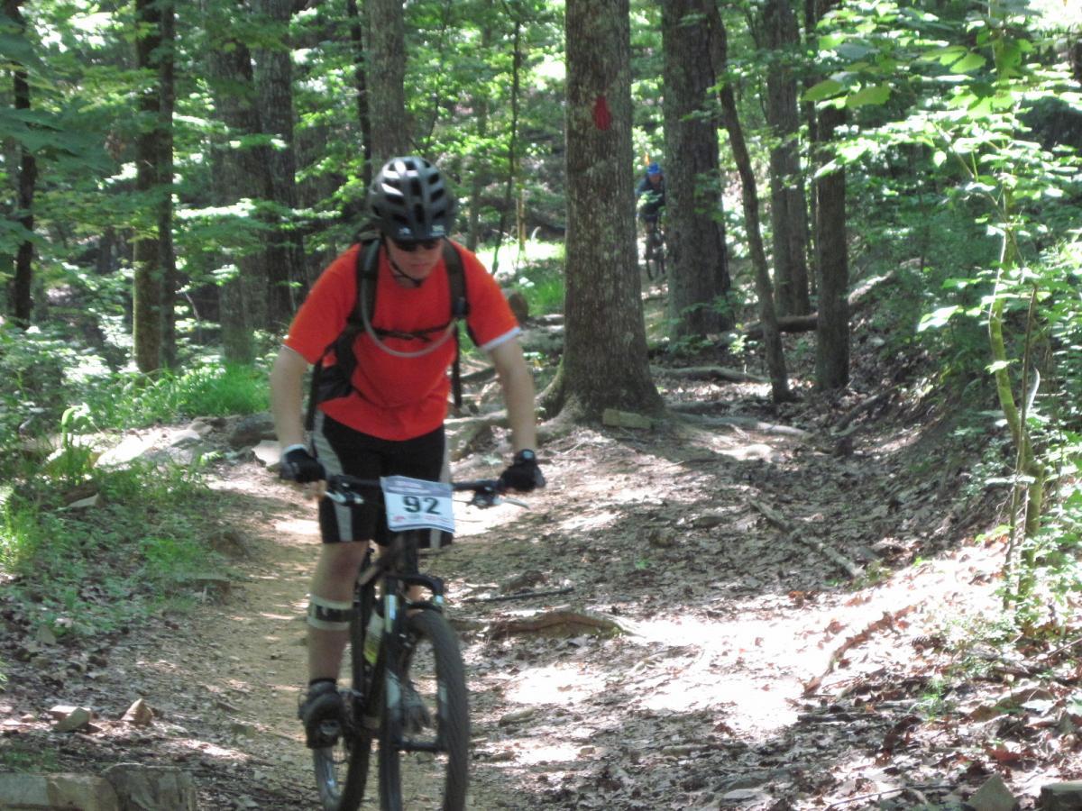 A mountain biker in a bright orange shirt rides along a narrow dirt trail in a wooded area, surrounded by green trees and foliage. The biker is focused on the path ahead, wearing a helmet and sunglasses, with a race number displayed on the bike. In the background, additional cyclists can be seen navigating the trail. Oak Mountain State Park Bump Trail mountain bike trail.
