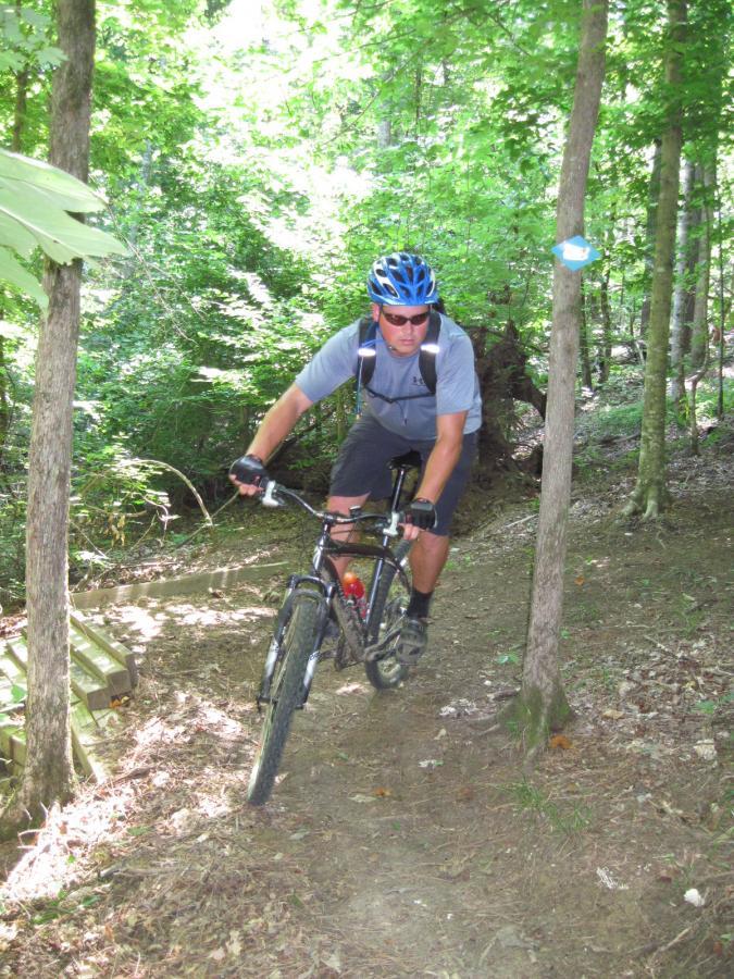 A cyclist riding a mountain bike on a narrow dirt trail surrounded by lush green trees. The rider is wearing a blue helmet and sunglasses, navigating the winding path in a forested area. Noxubee Crest mountain bike trail.