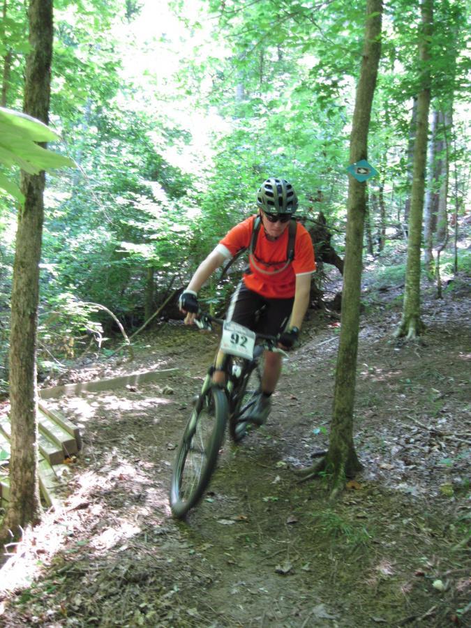 A mountain biker in an orange shirt and helmet navigates a dirt trail surrounded by trees. The cyclist wears a number 92 on their bike, and there are wooden planks visible in the path. The scene is bright and lush, showcasing a vibrant forest environment. Noxubee Crest mountain bike trail.