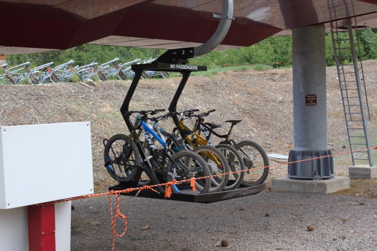 Four bicycles are positioned on a bike lift, designed to transport them up a mountain. A sign is visible reading "NO PASSENGERS." The lift is partially sheltered, and in the background, several bikes are parked on the ground. The area is surrounded by greenery and gravel. Northstar Bike Park mountain bike trail.