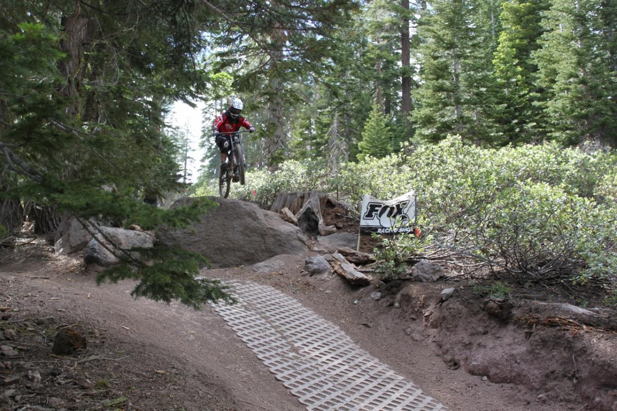 A mountain biker wearing a helmet and red jersey jumps off a rock on a dirt trail surrounded by trees. A banner for "Fox Racing Shox" is visible in the background, and a section of textured matting runs along the trail. Northstar Bike Park mountain bike trail.