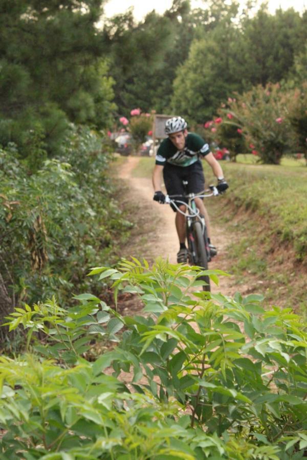 A person riding a mountain bike on a dirt path surrounded by greenery, with trees and flowering plants in the background. The cyclist is wearing a helmet and cycling attire, focused on navigating the trail. Mt Tabor Park mountain bike trail.
