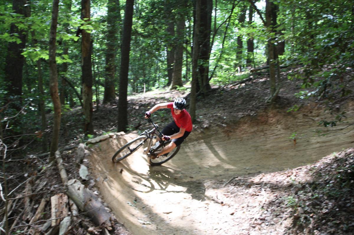 A person riding a mountain bike on a dirt trail surrounded by tall trees and lush greenery, leaning into a bend in the track. The scene captures the thrill of outdoor cycling in a natural environment. Warrior Creek mountain bike trail.
