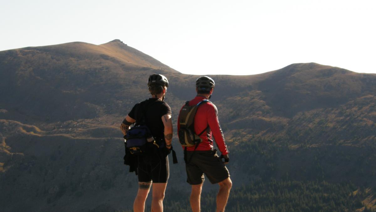 Two mountain bikers, one wearing a black shirt and the other in a red shirt, stand at the edge of a mountain overlook, gazing at the distant hills and landscape. They are equipped with helmets and backpacks, surrounded by a scenic backdrop of rolling mountains under a clear sky. Monarch Crest Trail mountain bike trail.