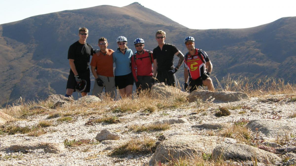 A group of six people standing on a rocky surface with a mountainous landscape in the background. They are dressed in casual outdoor clothing and biking gear, smiling as they pose for the photo under clear blue skies. The terrain is grassy with scattered rocks, suggesting a high-altitude hiking or biking location. Monarch Crest Trail mountain bike trail.