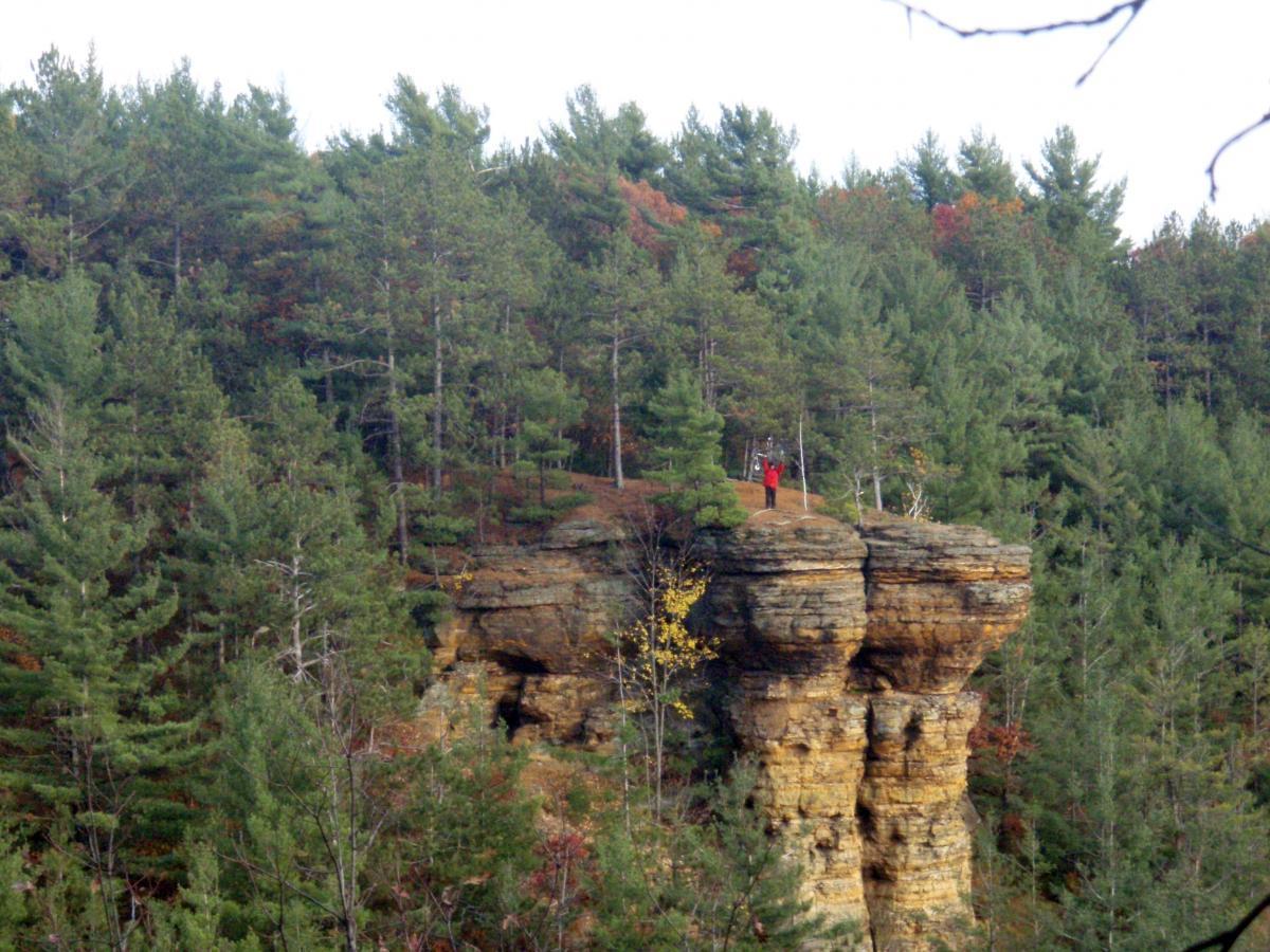 A person standing on the edge of a rocky cliff surrounded by dense green forest. The scene captures autumn colors in the foliage, with a cloudy sky overhead. Levis Mounds mountain bike trail.