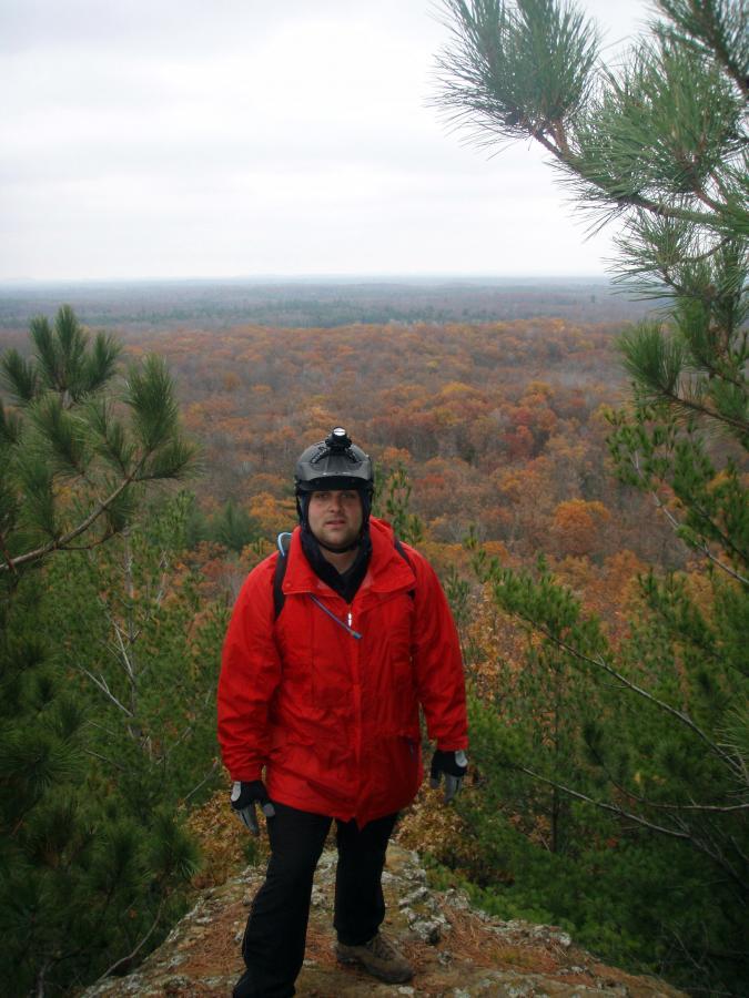 A person wearing a red jacket and a helmet stands on a rocky outcrop overlooking a colorful autumn landscape filled with trees. The sky is overcast, and the scene captures a mix of green pines and vibrant orange and yellow foliage in the distance. Levis Mounds mountain bike trail.