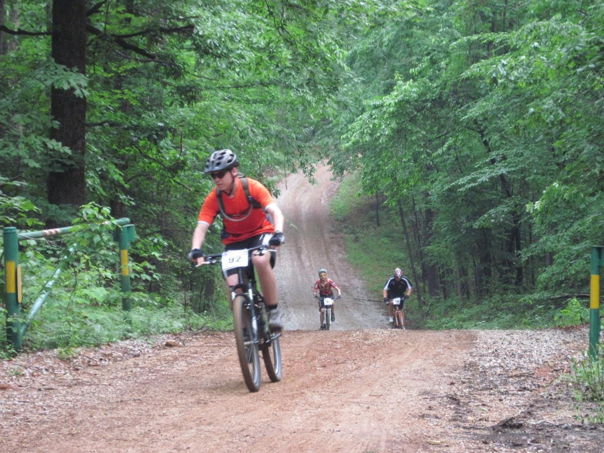 A group of three mountain bikers riding along a dirt path surrounded by lush greenery. One cyclist in the foreground is wearing an orange shirt and a helmet, while the other two are further behind, each wearing different colored jerseys. The atmosphere is damp, suggesting recent rain, with tall trees lining the trail. Noxubee Crest mountain bike trail.