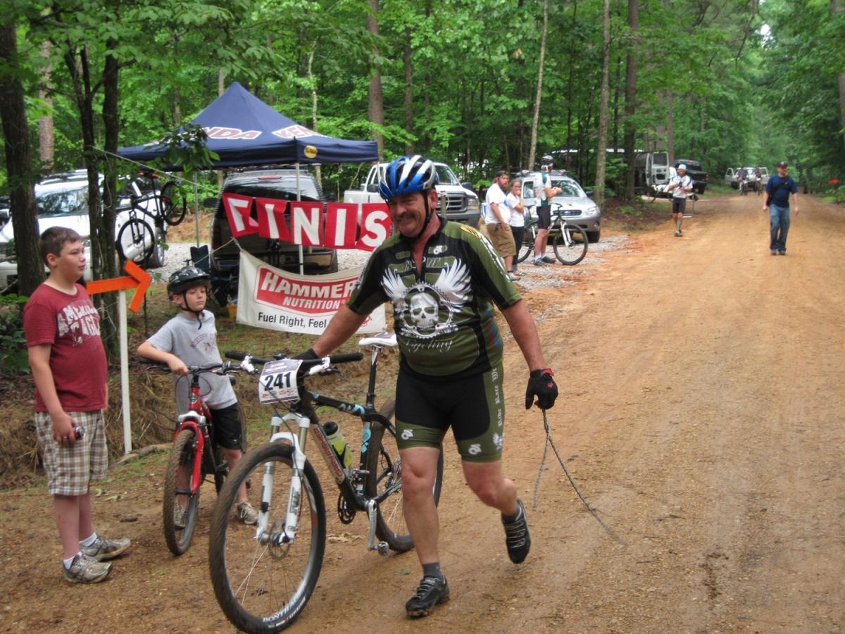 A male cyclist in a patterned jersey and helmet is walking a mountain bike along a dirt path, nearing a finish line marked by a banner. Two young boys are nearby on their bikes, watching the cyclist. The scene is set in a wooded area, with vehicles parked in the background. Noxubee Crest mountain bike trail.