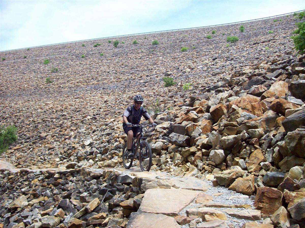 A mountain biker navigating a rocky trail on a hillside covered with loose stones and boulders, surrounded by patches of greenery and a cloudy sky above. Raccoon Mountain Trail Network mountain bike trail.