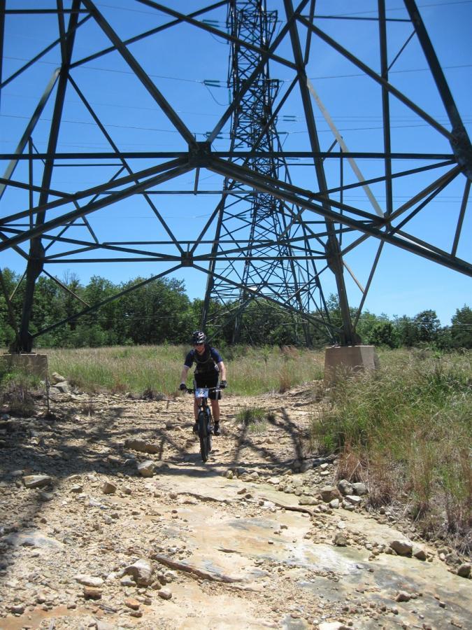 A mountain biker rides on a rocky trail beneath a large transmission tower, surrounded by tall grass and trees under a clear blue sky. Raccoon Mountain Trail Network mountain bike trail.