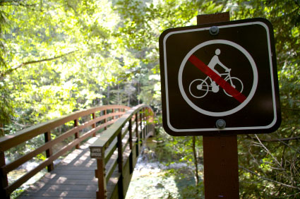 A wooden bridge surrounded by lush greenery, with a sign in the foreground indicating that bicycles are not allowed. The sign features a pictogram of a person riding a bicycle crossed out in red.