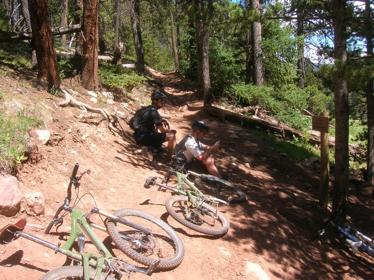 Two mountain bikers resting on a dirt trail surrounded by trees. Several bikes are laid on the ground nearby. One biker is sitting on the ground, while the other is sitting against a tree, appearing to check something on his phone. A trail sign can be seen in the background.  Reno / Flag / Bear / Deadman Loop mountain bike trail.