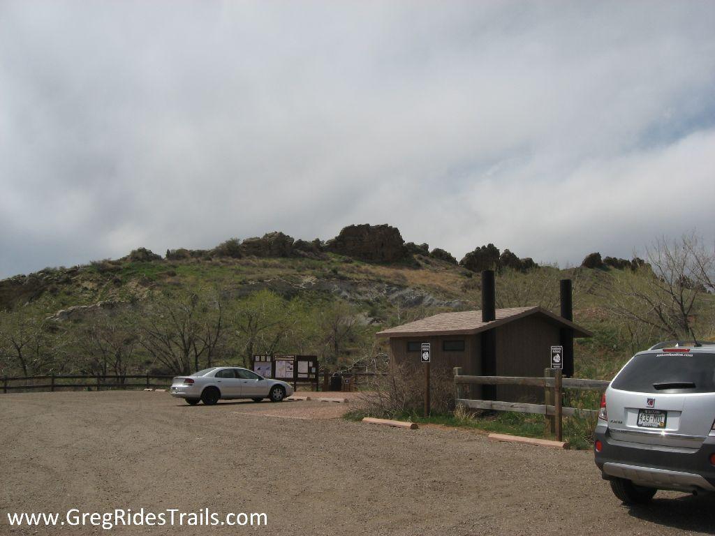 Parking area at the base of a rocky hillside, featuring a small restroom facility and several vehicles. The landscape is partly cloudy, with grassy and rocky terrain in the background. Devil's Backbone mountain bike trail.