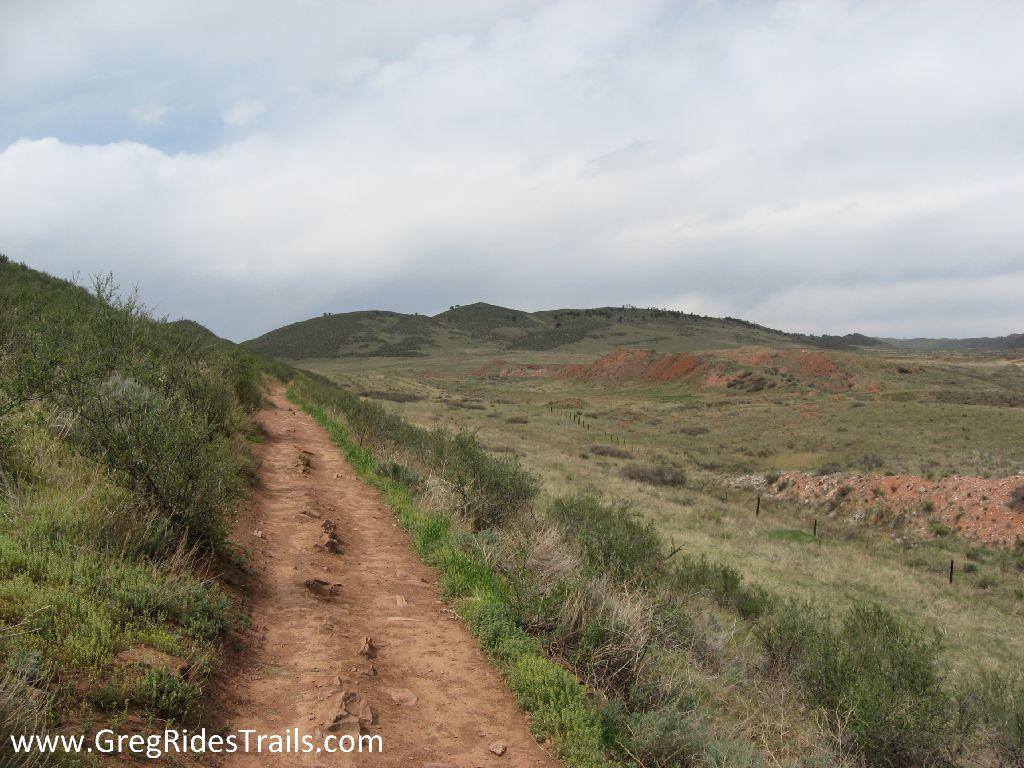 A dirt trail winding through green hills and grassy landscapes under a partly cloudy sky. The foreground shows a rough path bordered by shrubs, leading towards distant hilly terrain. Devil's Backbone mountain bike trail.