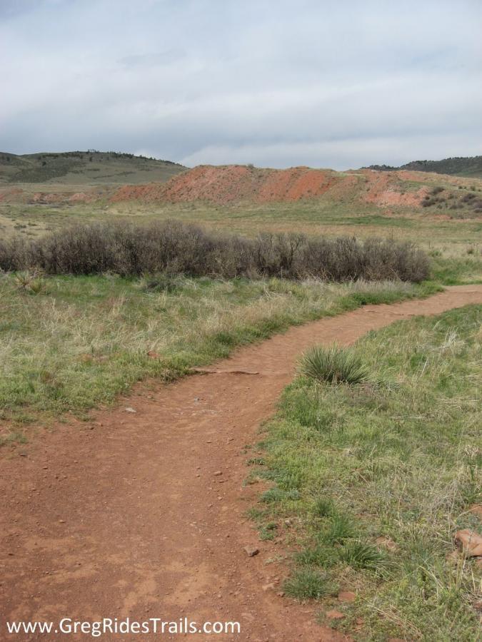 A winding dirt path meanders through an open grassy field, with patches of shrubbery on the sides. In the background, a reddish limestone formation rises against a cloudy sky, showcasing rolling hills and a serene natural landscape. Devil's Backbone mountain bike trail.