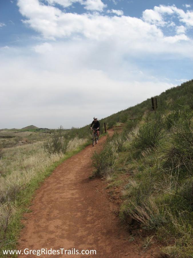 A cyclist riding on a dirt trail surrounded by green grass and shrubs, with a clear blue sky above. The path winds through a natural landscape, and hills can be seen in the distance. Devil's Backbone mountain bike trail.
