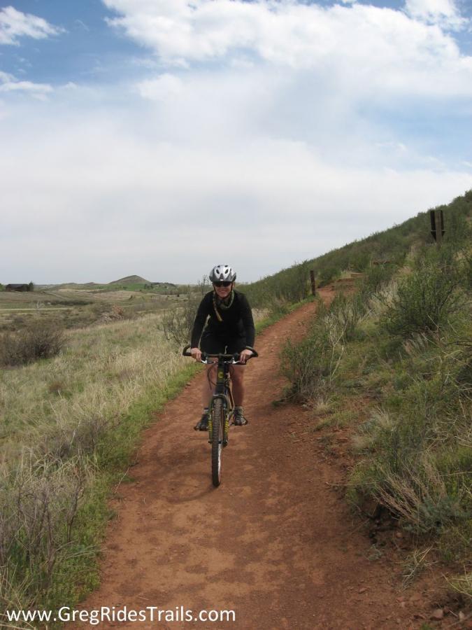 A cyclist riding a mountain bike on a dirt trail surrounded by grassy hills under a partly cloudy sky. Devil's Backbone mountain bike trail.