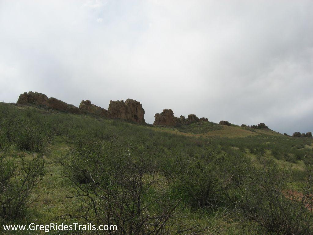 A scenic view of rocky formations rising above a grassy landscape under a cloudy sky. The foreground features low shrubs and greenery, while the rugged cliffs are visible in the background. Devil's Backbone mountain bike trail.
