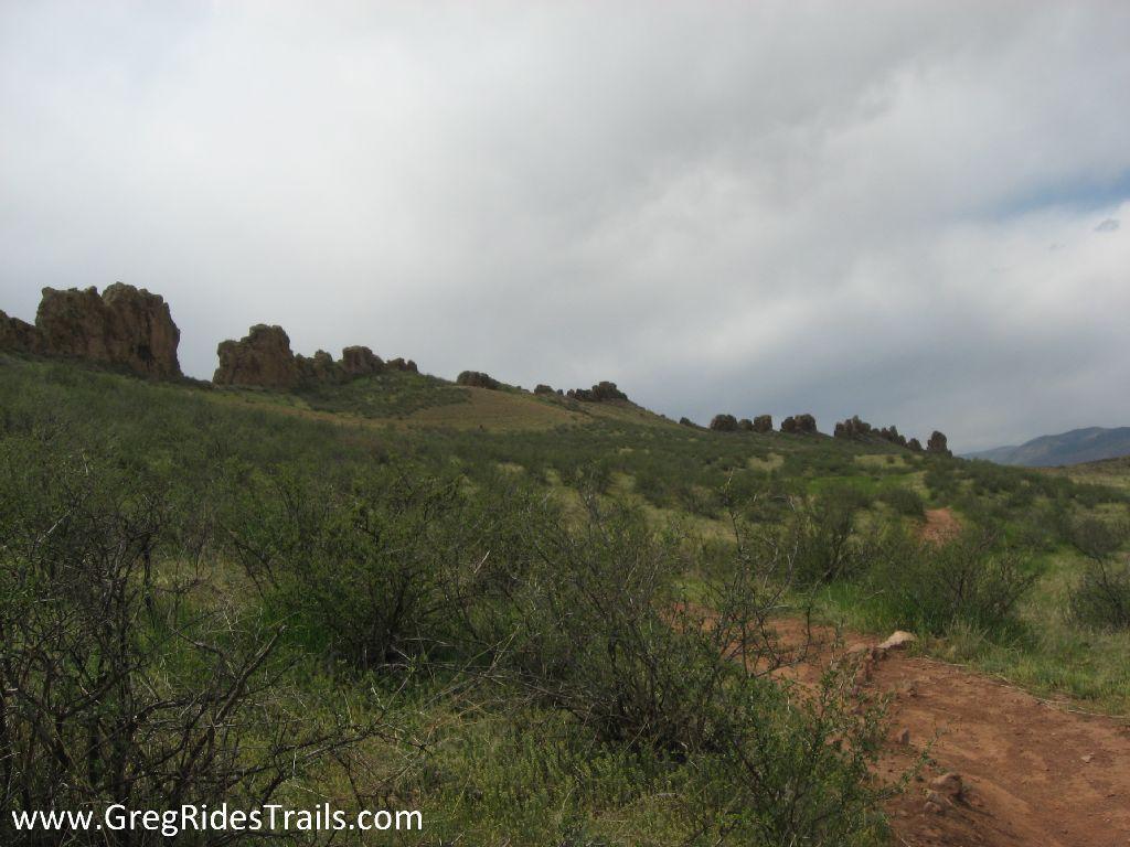 A scenic landscape featuring rocky formations on a hillside, with patches of green vegetation and a dirt trail winding through the foreground under a cloudy sky. Devil's Backbone mountain bike trail.