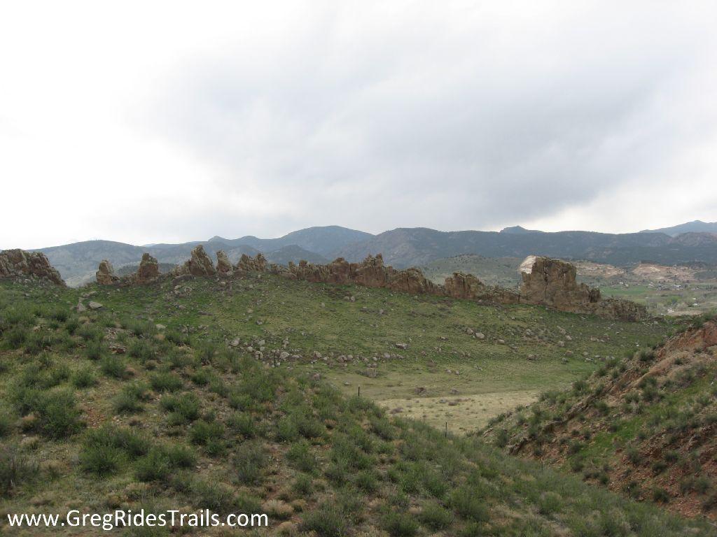 A panoramic view of a rocky landscape with green hills and underbrush, set against a cloudy sky and rolling mountains in the background. The scene features scattered rock formations and a mix of natural vegetation, creating a serene, outdoor atmosphere. Devil's Backbone mountain bike trail.