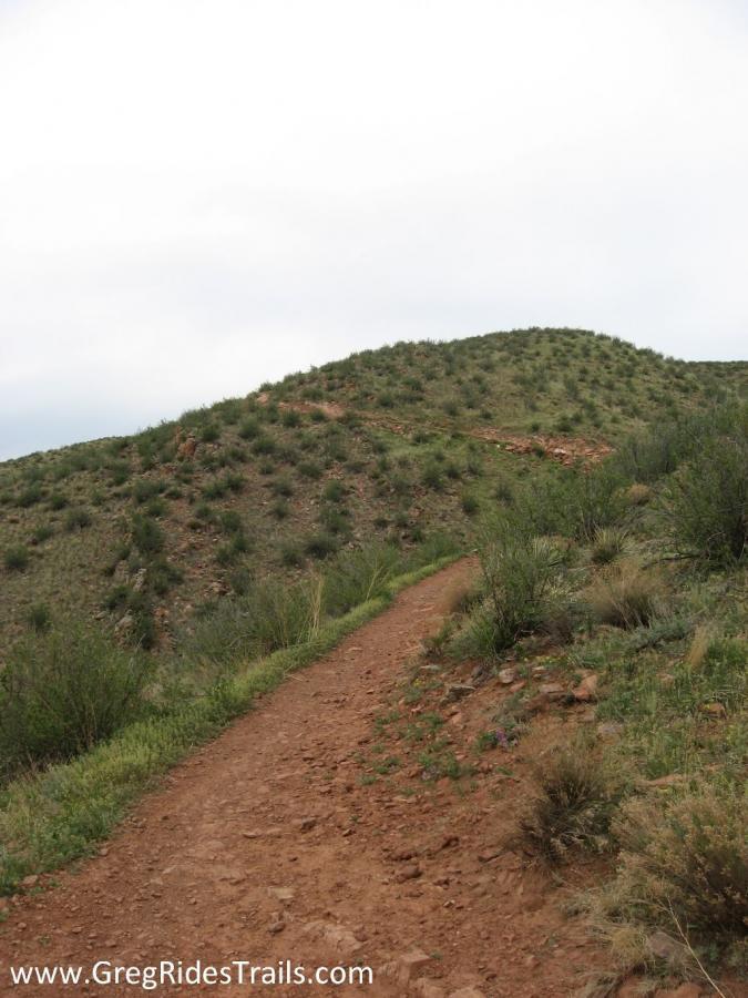 A winding dirt trail leads upwards through grassy and rocky terrain, flanked by low shrubs and plants, under a cloudy sky. The trail appears well-worn, suggesting it is frequently used for hiking or biking in a natural landscape. Devil's Backbone mountain bike trail.