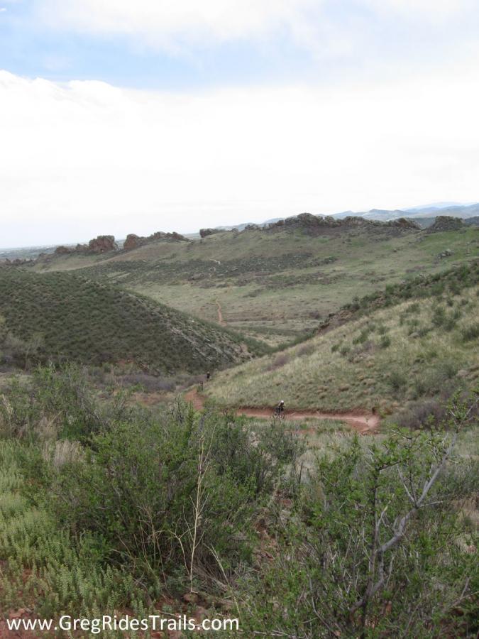 A panoramic view of a hilly landscape with green vegetation, featuring a dirt trail winding through the hills. The sky is partly cloudy, and a cyclist can be seen riding along the trail in the distance. Devil's Backbone mountain bike trail.