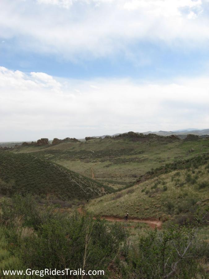 A scenic view of rolling green hills under a cloudy sky, featuring a dirt trail winding through the landscape. In the distance, a person on a bicycle can be seen making their way along the path, surrounded by lush vegetation and rocky formations. Devil's Backbone mountain bike trail.
