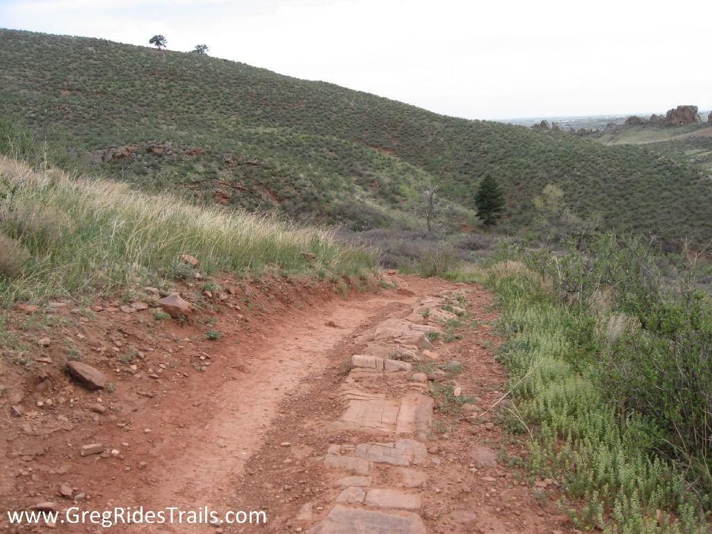 A winding dirt trail lined with rocks and bordered by green grasses and sparse shrubs, leading through rolling hills under a cloudy sky. Devil's Backbone mountain bike trail.
