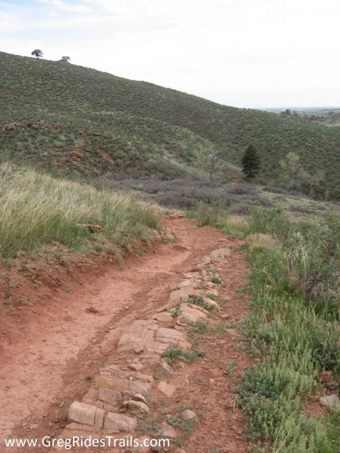 A winding dirt trail bordered by rocky sections, surrounded by green hills and sparse vegetation under a partly cloudy sky. Devil's Backbone mountain bike trail.