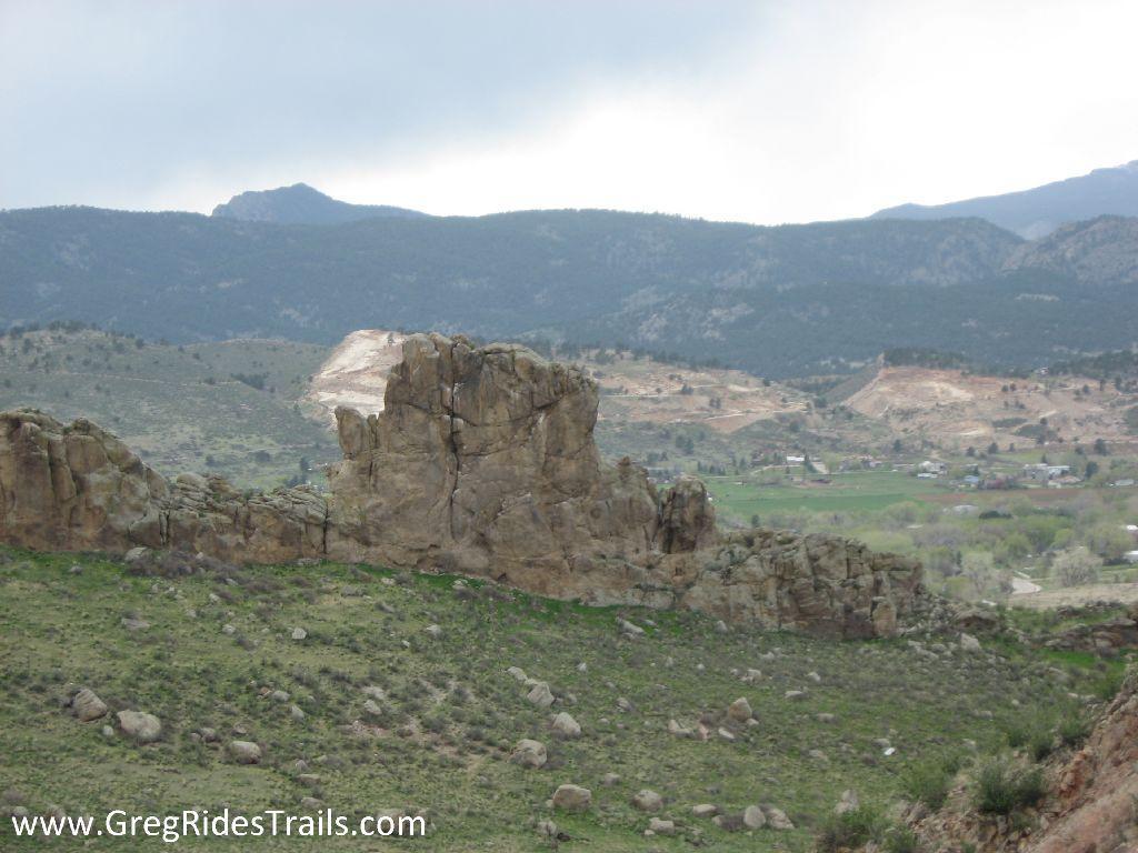 A rocky outcrop sits prominently in the foreground, surrounded by lush green grass and rolling hills. In the background, mountains rise under a cloudy sky, with a hint of a small settlement visible in the distance. The landscape features varied shades of green and earthy tones of the rocks, creating a picturesque natural scene. Devil's Backbone mountain bike trail.