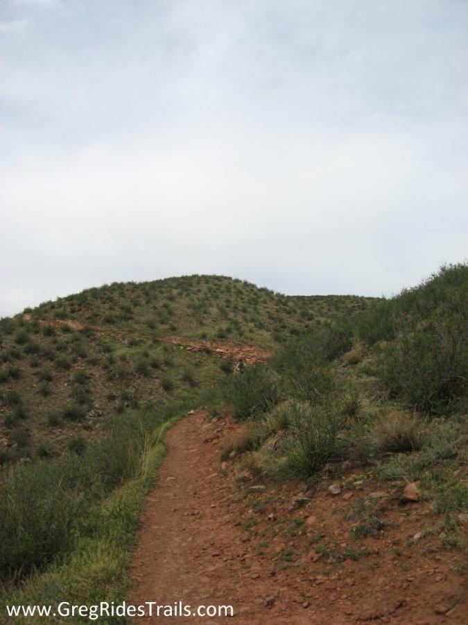 A winding dirt trail leads up a green, hilly landscape under a light cloudy sky. Sparse vegetation and small shrubs line the path, creating a natural setting for hiking or outdoor activities. Devil's Backbone mountain bike trail.