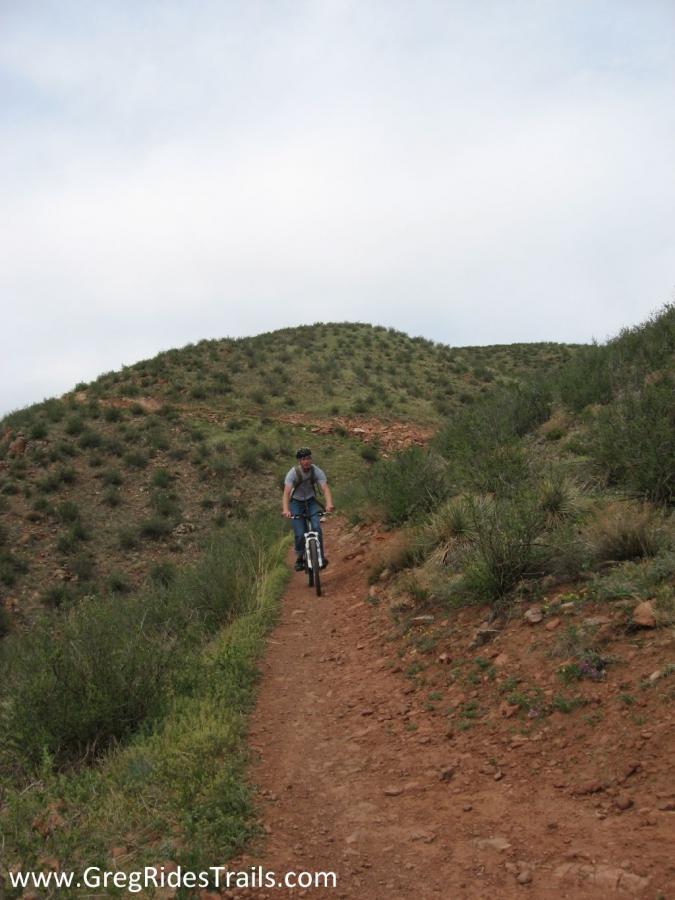 A cyclist riding a mountain bike on a dirt trail surrounded by greenery and rolling hills under a cloudy sky. The path winds up towards a hillside covered in low vegetation. Devil's Backbone mountain bike trail.