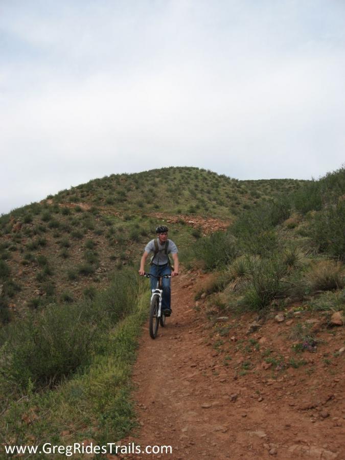 A cyclist riding a mountain bike on a dirt trail surrounded by greenery and hills under a cloudy sky. Devil's Backbone mountain bike trail.