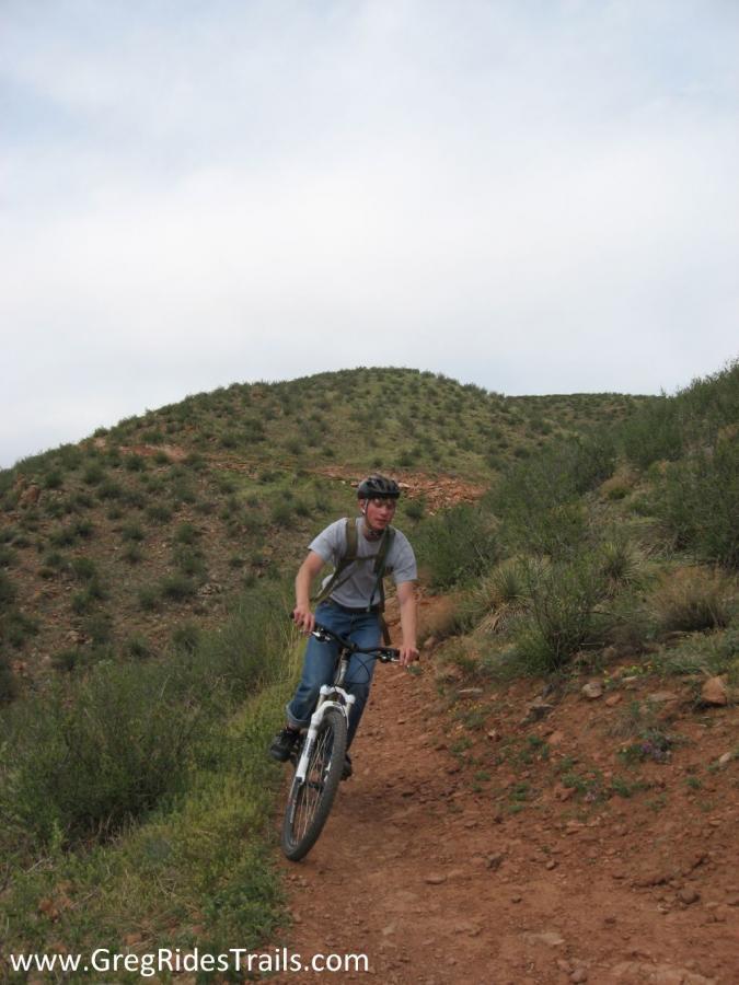 A person riding a mountain bike on a dirt trail surrounded by green vegetation and rolling hills under a cloudy sky. The cyclist is wearing a helmet and casual clothing, maintaining a focused posture as they navigate the terrain. Devil's Backbone mountain bike trail.