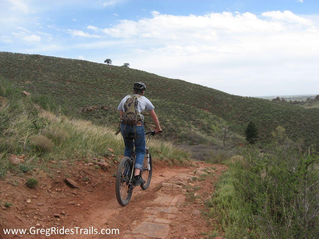 A person riding a mountain bike on a dirt trail surrounded by lush green hills and a blue sky with scattered clouds. The cyclist is wearing a helmet and a backpack, facing away from the camera as they navigate the trail. Devil's Backbone mountain bike trail.