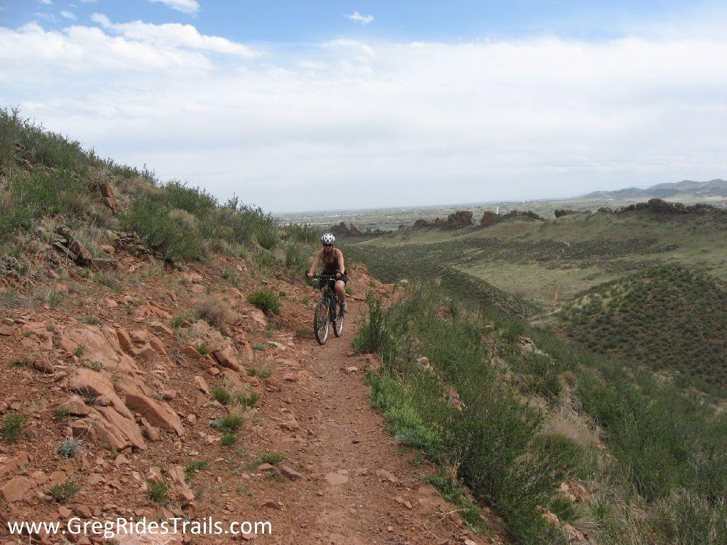A mountain biker navigating a rocky trail surrounded by green hills and an expansive sky in the background. Devil's Backbone mountain bike trail.
